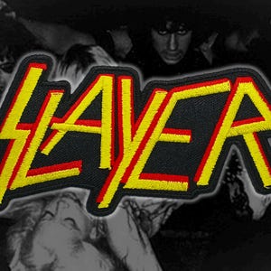 May include: Embroidered patch featuring the word "SLAYER" in yellow and red on a black background. The letters have a lightning bolt design. The patch is likely for a jacket or bag, representing a heavy metal band.