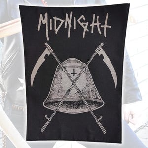 Peut inclure: Un écusson rectangulaire en tissu noir avec un motif sérigraphié blanc représentant une cloche ornée d'une croix inversée et deux faux croisées. Le mot MIDNIGHT est imprimé dans une police blanche stylisée au-dessus de l'illustration centrale.