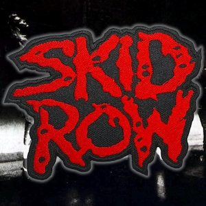 May include: A black patch with the red text "SKID ROW" in a stylized font. The patch has a black border and is set against a dark, blurred background. The patch is likely for a band.