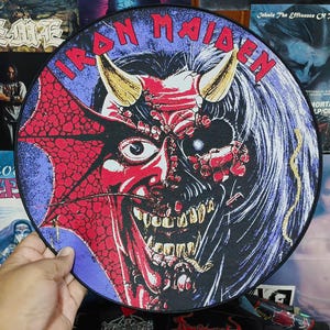 Op de afbeelding: Zwarte Iron Maiden LP met een demonisch gezicht in rood, wit en goud. De tekst "IRON MAIDEN" staat in het rood bovenaan. De LP wordt door een hand vastgehouden.