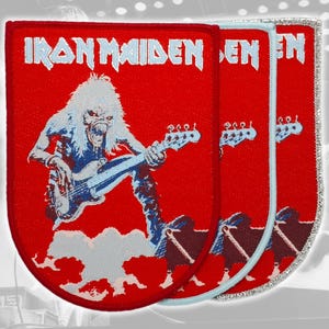Könnte beinhalten: Drei rechteckige gestickte Aufnäher mit abgerundeten unteren Ecken, rotem Hintergrund und dem weißen Schriftzug IRON MAIDEN. Jeder Aufnäher zeigt eine langhaarige Figur, die eine Bassgitarre über weißen Wolken spielt.
