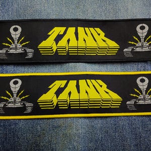 May include: Two rectangular patches with the word "TANK" in yellow, block-style letters. Each patch features a stylized tank graphic on each end. One patch has a black border, and the other has a yellow border.