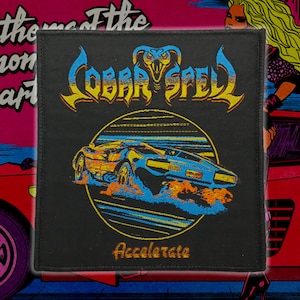 Puede incluir: Parche cuadrado negro con la inscripción "COBRA SPELL" en amarillo y azul, una cabeza de cobra sobre un coche estilizado y la palabra "Accelerate" debajo. Con borde cosido.