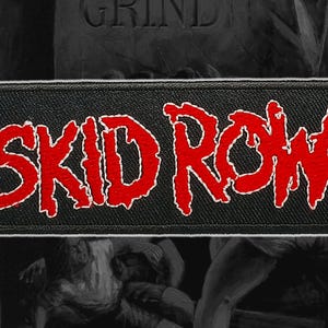 Puede incluir: Un parche rectangular negro con las palabras "SKID ROW" en rojo, delineadas en blanco. El texto está en negrita y estilizado. El parche es probablemente para ropa o accesorios. El fondo es oscuro.