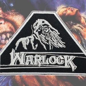 May include: Black and white embroidered patch with the word "WARLOCK" and a stylized creature design. The patch has a black background with a white border and is in a pentagon shape. The background features fantasy art.