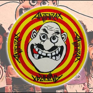 May include: A circular embroidered patch featuring a central cartoon face with a mustache and wide grin, surrounded by concentric red and yellow rings. The word ANTHRAX is printed in black lettering at the top, bottom, and sides of the border.