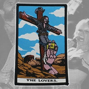 May include: Embroidered patch featuring a stylized illustration of a figure on a cross, with a second figure at the base. The background includes a blue sky and desert landscape. The text "THE LOVERS" is at the bottom.