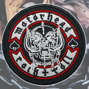 Puede incluir: Un parche bordado circular con fondo negro y borde blanco y rojo. El centro muestra un gráfico de calavera Snaggletooth blanca con cadenas. El texto Motorhead está arqueado arriba y el texto England está arqueado en la parte inferior.