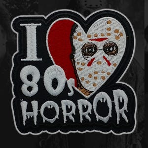 Könnte beinhalten: Gestickter Aufnäher mit dem Text "I 80s HORROR". Das Design zeigt eine Herzform mit geteiltem Design, eine Seite rot und die andere eine weiße Hockeymaske mit braunen und roten Details. Der Aufnäher hat einen schwarzen Rand.