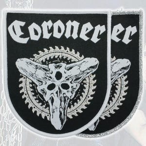 Könnte beinhalten: Zwei schwarze Stoffaufnäher mit weiß bestickten Rändern. Jeder Aufnäher zeigt das Wort Coroner in einer gotischen Schrift über einer silbernen, kreisförmigen Zahnradgrafik mit einem stilisierten, dreiäugigen Totenkopf-Design.