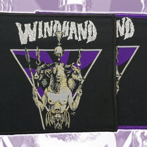 Pode incluir: Um emblema quadrado bordado em preto com a palavra WINDHAND em texto prateado sobre um gráfico de uma figura de cabra de três cabeças centrada num triângulo roxo. O emblema tem uma borda preta espessa e detalhes em fios prateados e roxos.