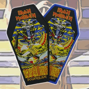 Puede incluir: Dos parches bordados con forma de ataúd que presentan el logotipo de Iron Maiden en texto rojo y amarillo. El diseño muestra una figura similar a un faraón en azul, amarillo y negro, con la palabra Powerslave impresa en amarillo en la parte inferior.