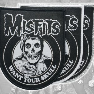 Może przedstawiać: Trzy czarno-białe haftowane naszywki z logo Misfits u góry i grafiką czaszki pośrodku. Napis WANT YOUR SKULL znajduje się w kolistym łuku pod czaszką. Każda naszywka posiada wykończenie krawędzi o innym charakterze.