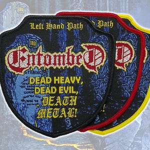 May include: Three shield-shaped embroidered patches featuring the text Left Hand Path, Entombed, and Dead Heavy, Dead Evil, Death Metal! The patches have black, red, and yellow borders respectively, with blue and black graphic backgrounds.