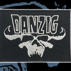 May include: Embroidered black patch featuring the word "DANZIG" in white, stylized lettering above a white skull design. The patch has a white border and is likely for clothing or accessories.