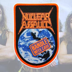 May include: A rectangular embroidered patch with rounded corners and a bright orange border. It features the text NUCLEAR ASSAULT in orange, a graphic of the Earth, and a tilted orange rectangular label with the text HANDLE WITH CARE.
