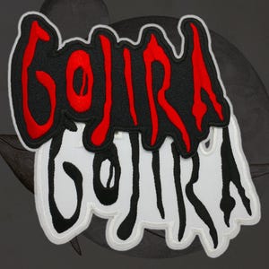 May include: Embroidered patch featuring the word "GOJIRA" in red, stylized lettering over a black background, with a white and black version below. The patch has a white border and is likely for clothing or accessories.