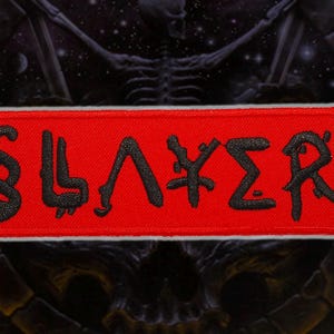 Könnte beinhalten: Rechteckiger Aufnäher mit dem Wort „SLAYER“ in schwarzer, stilisierter Schrift auf rotem Grund. Der Aufnäher hat einen weißen Rand. Der Hintergrund ist dunkel und strukturiert.