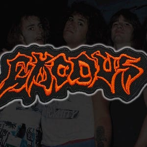 May include: Embroidered patch with the word "BROODS" in orange, stylized lettering on a black background. The patch is rectangular with a wavy border. The background features a blurred image of a group of people.
