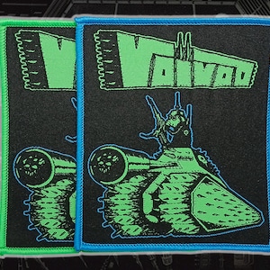 May include: Two rectangular embroidered patches featuring the word VOIVOD in bright green text above a green graphic of a tank with a figure on top. One patch has a bright green border, and the other has a bright blue border on a black background.