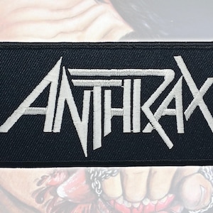 Può includere: Una toppa rettangolare nera ricamata con la parola ANTHRAX in lettere maiuscole bianche. Il testo è stilizzato con linee angolari nette e una barra orizzontale che si estende dalla parte superiore della lettera T sopra le lettere H, R e A.