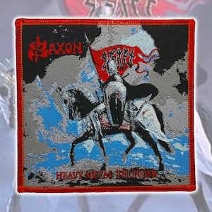 May include: A square embroidered patch with a red border featuring a graphic of a knight on horseback holding a red flag. The text Saxon appears in red at the top left, and the text Heavy Metal Thunder is printed in red at the bottom center.