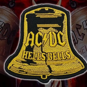 May include: Embroidered patch featuring the words "AC/DC" above "HELLS BELLS" in yellow thread on a black bell-shaped background. The patch has a black border and is set against a blurred background of bells.