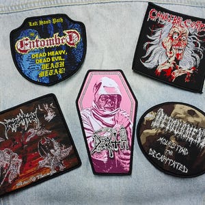 May include: Five embroidered fabric patches featuring heavy metal band logos and artwork. Designs include Entombed, Cannibal Corpse, Immolation, Death in a pink coffin shape, and Devourment. Each patch has a black border and detailed graphics.