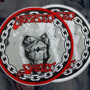 May include: Two round patches with a skull graphic. The patches feature the words "Morbid Saint" in red, with a chain border. The skull is gray and black, set against a gray background.