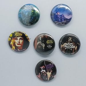 May include: Six circular pin-back buttons featuring King Diamond album artwork. Designs include Abigail, Them, Fatal Portrait, The Dark Sides, The Family Ghost, and a portrait of the musician with the King Diamond logo on each button.