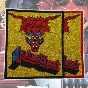 May include: Two square patches with a gold glitter background. The patches feature a red and blue dragon design above the text "Judas Priest" in a red and blue font. One patch has a black border, the other has a red border.