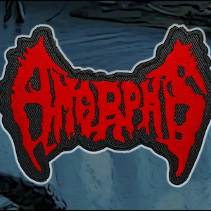 May include: A rectangular embroidered patch featuring the word Amorphis in a stylized, jagged red font. The lettering is set against a black background with a white border outline, creating a high-contrast design with sharp, angular edges.