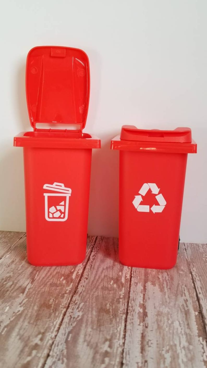 May include: Two red plastic trash cans with lids. One has a white recycling symbol and the other has a white trash can symbol.