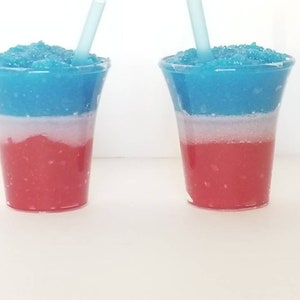 May include: Two clear plastic cups filled with a layered blue, white, and red slushie.  Each cup has a blue straw sticking out of the top.