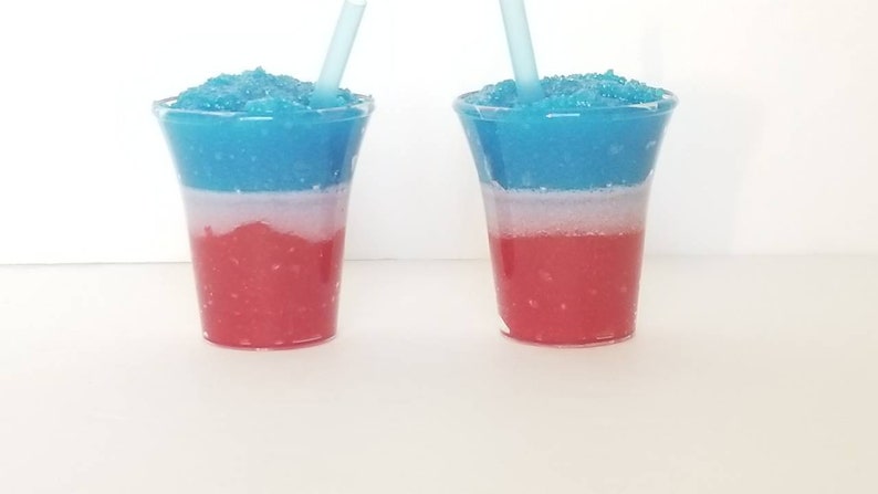 May include: Two clear plastic cups filled with a layered blue, white, and red slushie.  Each cup has a blue straw sticking out of the top.