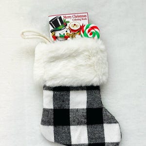 May include: A black and white buffalo plaid Christmas stocking with a white faux fur cuff. The stocking is filled with a small coloring book that says "Merry Christmas Coloring Book" and a red and green candy cane.