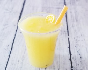 May include: A clear plastic cup filled with a bright yellow blended beverage. A yellow straw is inserted into the drink, and a small, decorative yellow and white citrus slice garnish rests on the surface of the liquid.