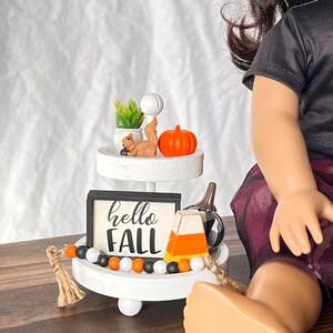 K&ouml;nnte beinhalten: Ein wei&szlig;er Etagere mit einem schwarz-wei&szlig;en Schild mit der Aufschrift "hello fall" und einem kleinen orangefarbenen K&uuml;rbis, einer Eichh&ouml;rnchenfigur, einer Mais-S&uuml;&szlig;warenfigur und einer schwarz-wei&szlig;en Perlenkette.