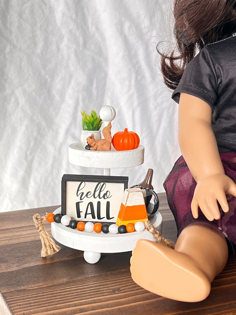 K&ouml;nnte beinhalten: Ein wei&szlig;er Etagere mit einem schwarz-wei&szlig;en Schild mit der Aufschrift "hello fall" und einem kleinen orangefarbenen K&uuml;rbis, einer Eichh&ouml;rnchenfigur, einer Mais-S&uuml;&szlig;warenfigur und einer schwarz-wei&szlig;en Perlenkette.
