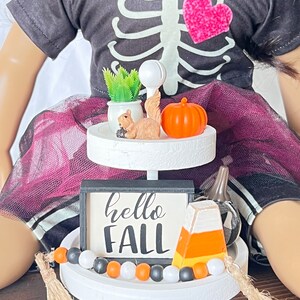 K&ouml;nnte beinhalten: Ein zweistufiger wei&szlig;er Etagere mit Herbstthema. Die obere Stufe hat eine kleine Topfpflanze, eine Eichh&ouml;rnchenfigur und einen kleinen orangefarbenen K&uuml;rbis. Die untere Stufe hat ein schwarz-wei&szlig;es Schild mit der Aufschrift "hello fall", eine Zuckermaisfigur und eine schwarz-wei&szlig;e Perlenkette.