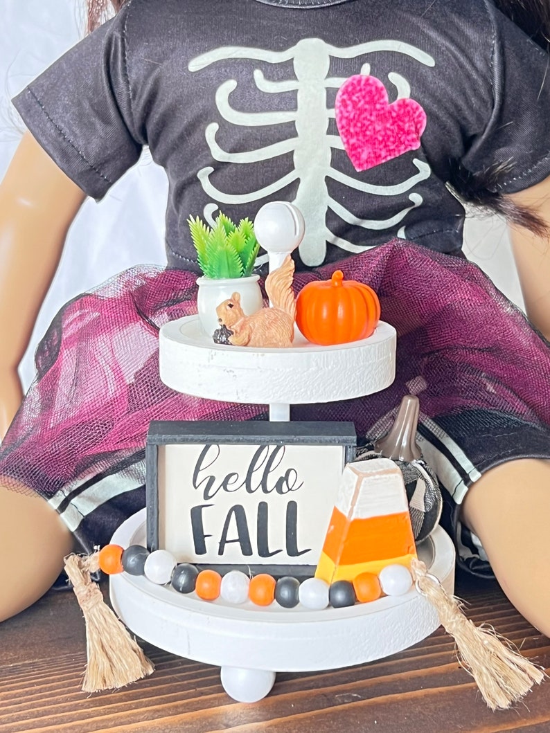 K&ouml;nnte beinhalten: Ein zweistufiger wei&szlig;er Etagere mit Herbstthema. Die obere Stufe hat eine kleine Topfpflanze, eine Eichh&ouml;rnchenfigur und einen kleinen orangefarbenen K&uuml;rbis. Die untere Stufe hat ein schwarz-wei&szlig;es Schild mit der Aufschrift "hello fall", eine Zuckermaisfigur und eine schwarz-wei&szlig;e Perlenkette.