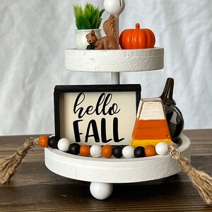 K&ouml;nnte beinhalten: Ein zweistufiger wei&szlig;er Etagere, dekoriert mit Herbst-Artikeln, darunter ein kleiner K&uuml;rbis, eine Eichh&ouml;rnchenfigur, eine Topfpflanze, ein Schild mit der Aufschrift "hello fall", ein Maisgeb&auml;ck und ein schwarz-wei&szlig; kariertes Stoffst&uuml;ck. Die Etagere ist mit einer schwarzen, wei&szlig;en und orangefarbenen Perlenkette verziert.