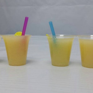 May include: Four clear plastic cups filled with a yellow liquid, each with a different colored straw. The straws are pink, blue, green, and orange.