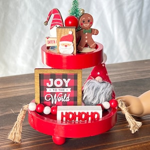 K&ouml;nnte beinhalten: Ein dreistufiger roter Etagere, dekoriert mit Weihnachtsfiguren, darunter ein Lebkuchenmann, ein Zwerg und ein Weihnachtsmann. Die Etagere hat auch ein Schild mit der Aufschrift "Joy to the World" und ein Schild mit der Aufschrift "HOHOHO".