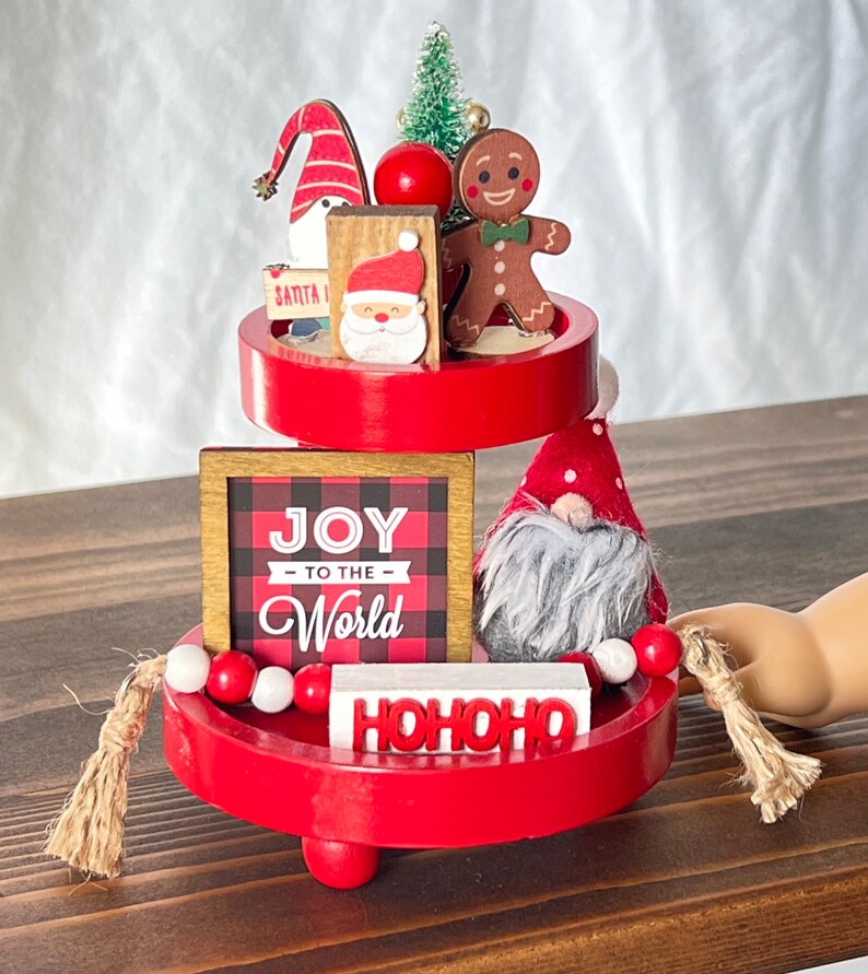 K&ouml;nnte beinhalten: Ein dreistufiger roter Etagere, dekoriert mit Weihnachtsfiguren, darunter ein Lebkuchenmann, ein Zwerg und ein Weihnachtsmann. Die Etagere hat auch ein Schild mit der Aufschrift "Joy to the World" und ein Schild mit der Aufschrift "HOHOHO".