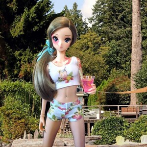 May include: A doll wearing a white crop top with a lemonade print and blue floral shorts. The doll is holding a pink drink with a straw. The doll has long brown hair and blue eyes.