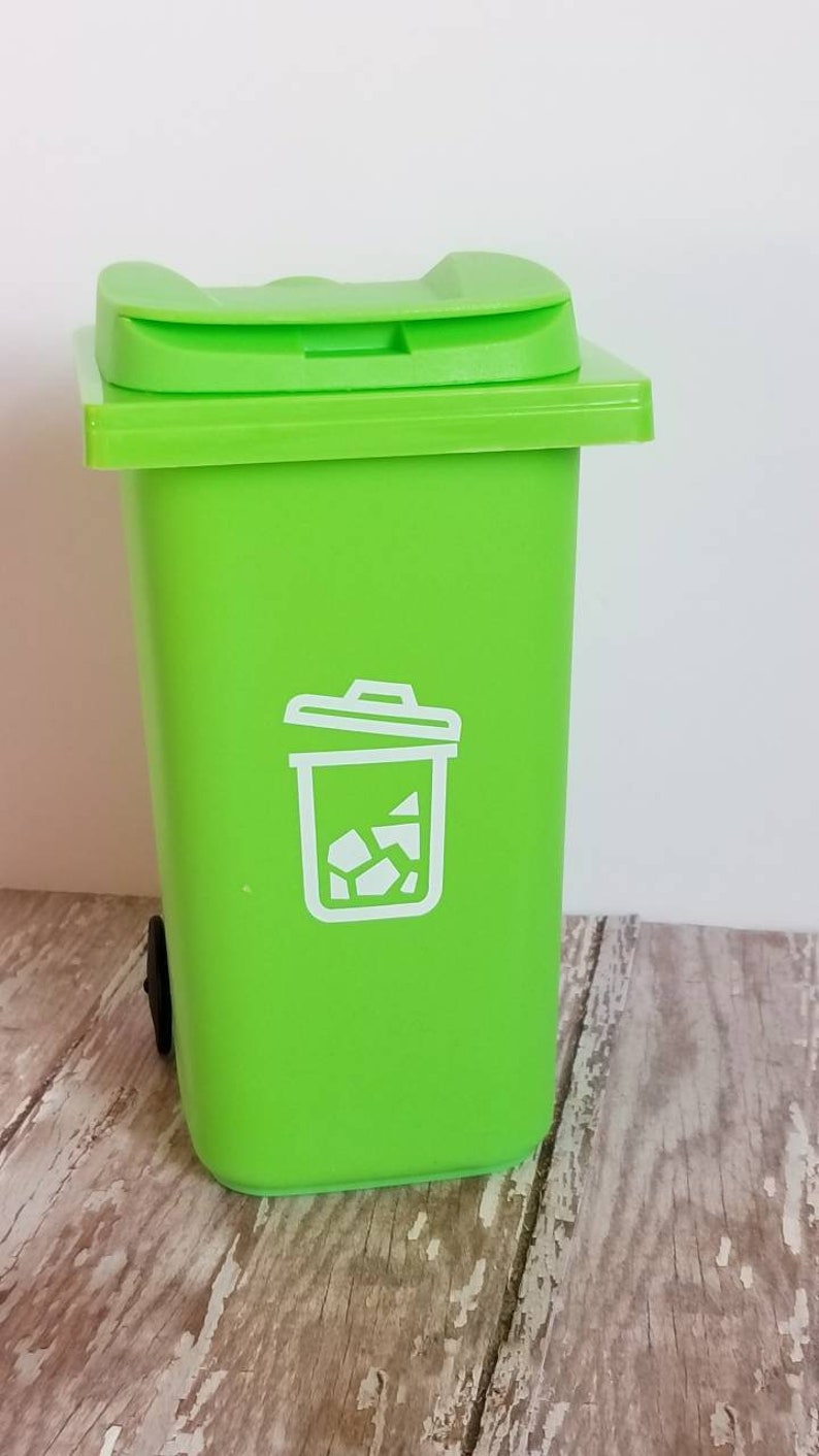 May include: A bright green plastic trash can with a white recycling symbol on the front. The can has a lid and a small wheel on the bottom.