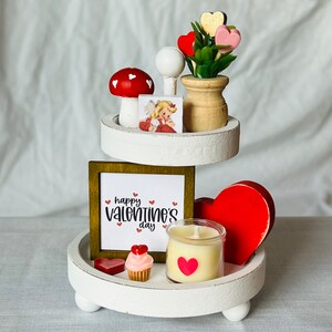 K&ouml;nnte beinhalten: Wei&szlig;er dreistufiger Tablett, dekoriert f&uuml;r Valentinstag mit einem roten Herzen, einem rot-wei&szlig;en Pilz, einer kleinen Kerze, einem Cupcake und einem Schild mit der Aufschrift "Happy Valentine's Day".