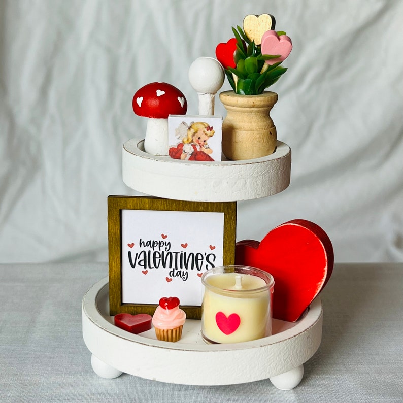K&ouml;nnte beinhalten: Wei&szlig;er dreistufiger Tablett, dekoriert f&uuml;r Valentinstag mit einem roten Herzen, einem rot-wei&szlig;en Pilz, einer kleinen Kerze, einem Cupcake und einem Schild mit der Aufschrift "Happy Valentine's Day".