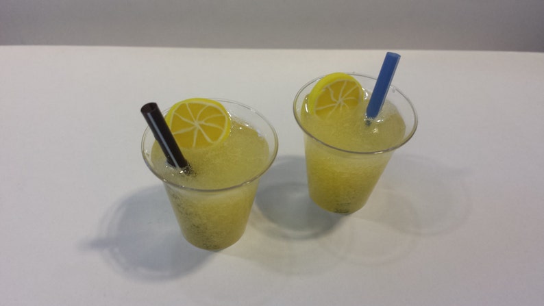 May include: Two clear plastic cups filled with a yellow liquid, topped with lemon slices and straws. One cup has a blue straw and the other has a black straw.